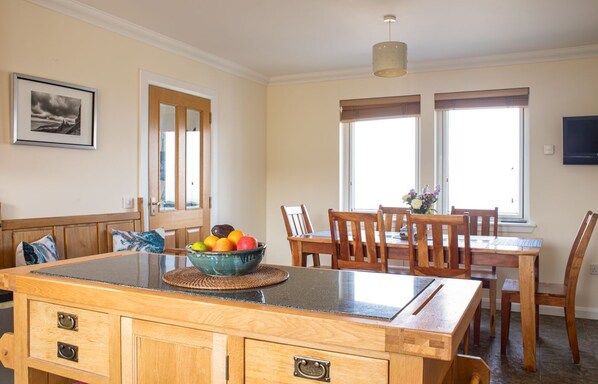 Een keuken met een houten keukeneiland, een eettafel met stoelen, een fruitschaal en een raam met jaloezieën.