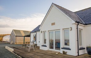 A white building with a sloped roof, large windows, and a wooden deck with a bench. - Solas Alainn -  a detached house that sleeps 6 guests  in 3 bedrooms (Staffin)