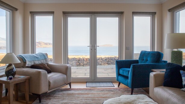 A living room with a beige sofa, a blue armchair, a lamp, and a view of the sea through large glass doors. - Solas Alainn -  a detached house that sleeps 6 guests  in 3 bedrooms (Staffin)