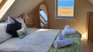 A bedroom with a bed, pillows, a mirror, a window with a beach view, and a pair of towels.
