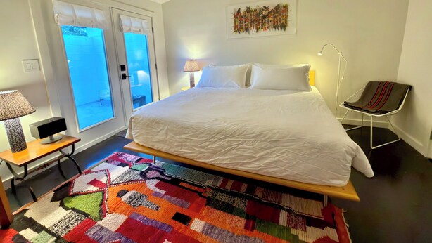 Um quarto com cama, tapete colorido, poltrona, luminária e janela com vista.