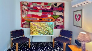 Quarto com uma grande tapeçaria colorida, uma televisão de tela plana, duas cadeiras, um tapete estampado e um quadro na parede.