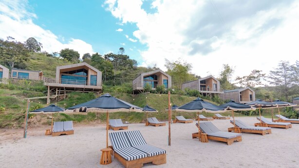 A beach resort with wooden beach chairs and umbrellas, wooden cabins with balconies, and a clear sky. - Okai by Bernalo Hotels (Guatape)