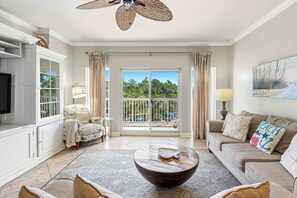 A living room with a large window, a ceiling fan, a flat-screen TV, and a sofa with decorative pillows. - 2BR condo with pool view & beach access (Miramar Beach)