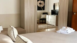 Una cama bien hecha con una colcha texturizada y almohadas decorativas, un espejo que refleja una habitación con un televisor y una silla, y una pila de toallas dobladas sobre la cama.