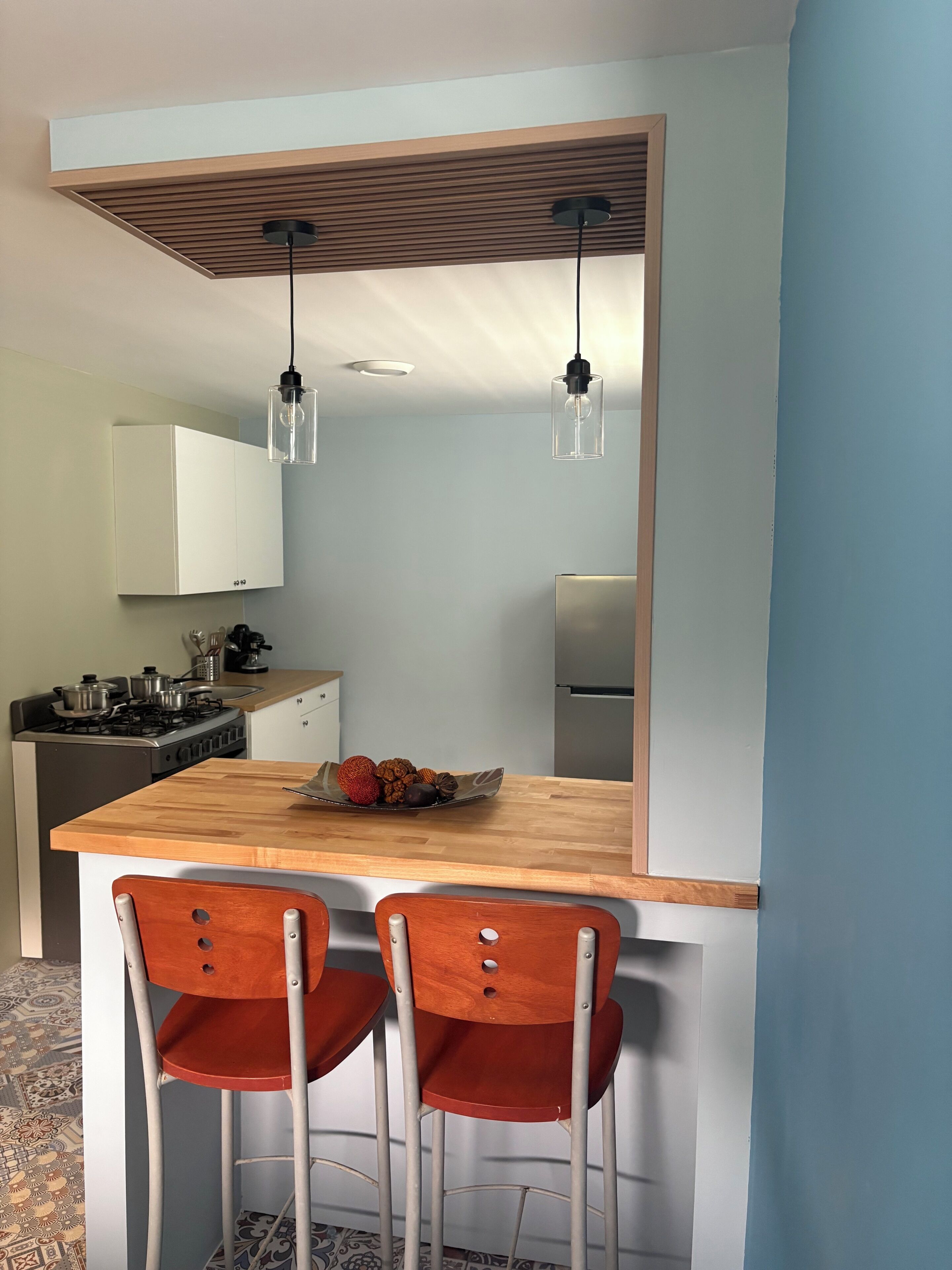 A modern kitchen with a wooden island, two red chairs, pendant lights, and a stainless steel refrigerator.
