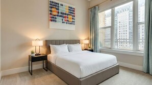 Quarto de hotel com cama, duas mesas de cabeceira com luminárias, uma janela grande com cortinas e um quadro abstrato colorido na parede.