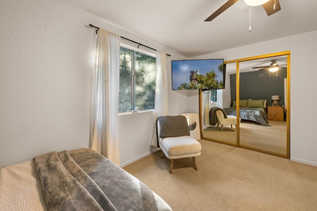 A bedroom with a bed, a chair, a window, a ceiling fan, and a mirror.