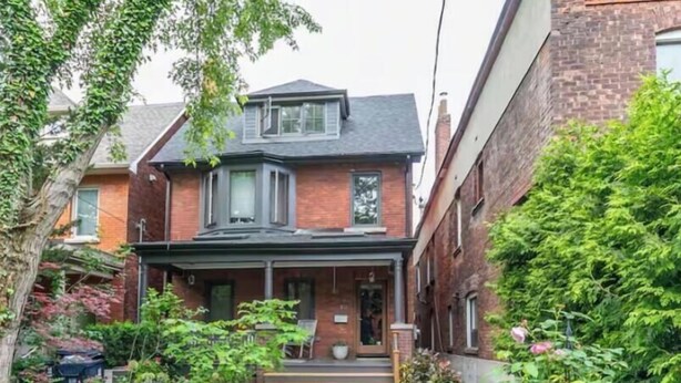Une maison à deux étages avec un porche à l’avant, une façade en briques et un jardin où poussent diverses plantes et fleurs.
