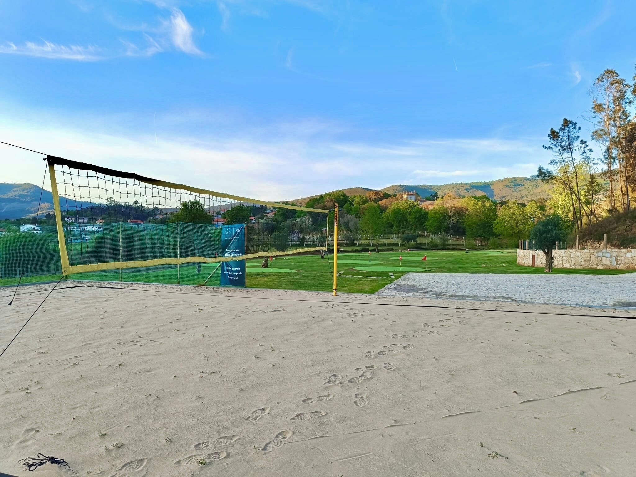 A volleyball court with a net, a sandy area, and a grassy field with trees and hills in the background.