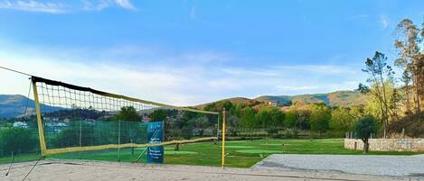 A volleyball court with a net, a sandy area, and a grassy field with trees and hills in the background.