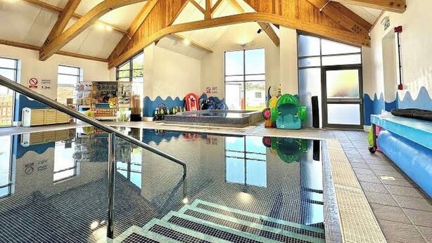 Una piscina coperta con soffitto in legno, barriera in vetro e area giochi per bambini sullo sfondo.