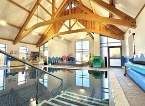 Una piscina interior amb sostre de fusta, una barrera de vidre i una zona de jocs per a nens al fons.