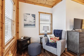 A cozy room with wooden walls, a grey sofa with a Union Jack pillow, a small table with a lamp, and a TV on a wooden cabinet.