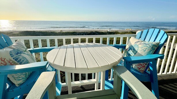 A deck with a round table and blue chairs, overlooking a beach.