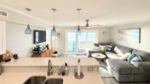 A modern kitchen with a central island, a dining area with a gray sofa, and a view of the ocean through the sliding glass doors.