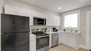 A modern kitchen with white cabinetry, a black refrigerator, stainless steel appliances, and a window with blinds. - Cozy Indy Retreat. Walk to Lucas Oil & Downtown. (Indianapolis)