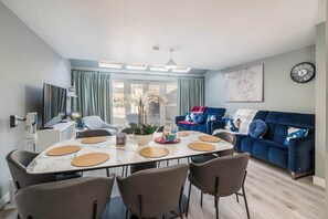 Ein modernes Wohnzimmer mit einem blauen Sofa, einem Esstisch mit Stühlen und einem großen Fenster mit Vorhängen.