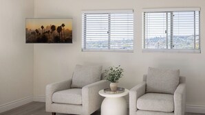 Uma sala de estar com dois poltronas, uma mesinha de apoio branca com uma planta e um quadro na parede.