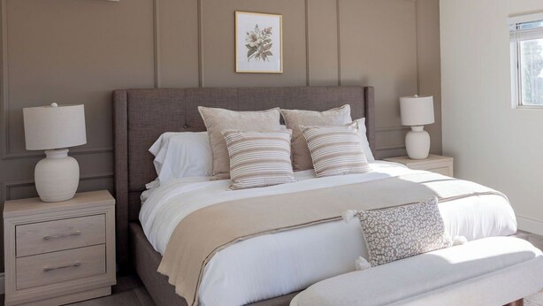 Um quarto com uma cama grande, duas mesas de cabeceira com luminárias, um banquinho e uma impressão botânica emoldurada na parede.