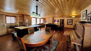 Eine geräumige Küche mit Holzausstattung, ein Essbereich mit großem Holztisch und ein Wohnzimmer mit Sofa und Fernseher.