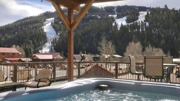 Ein Whirlpool mit einer Holzterrasse und Stühlen mit Blick auf eine verschneite Berglandschaft.