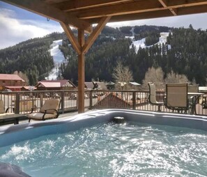 A hot tub with a wooden deck and chairs, overlooking a snowy mountain landscape. - Alpine Summit Lodge - Formally 4K Penthouse View (Red River)