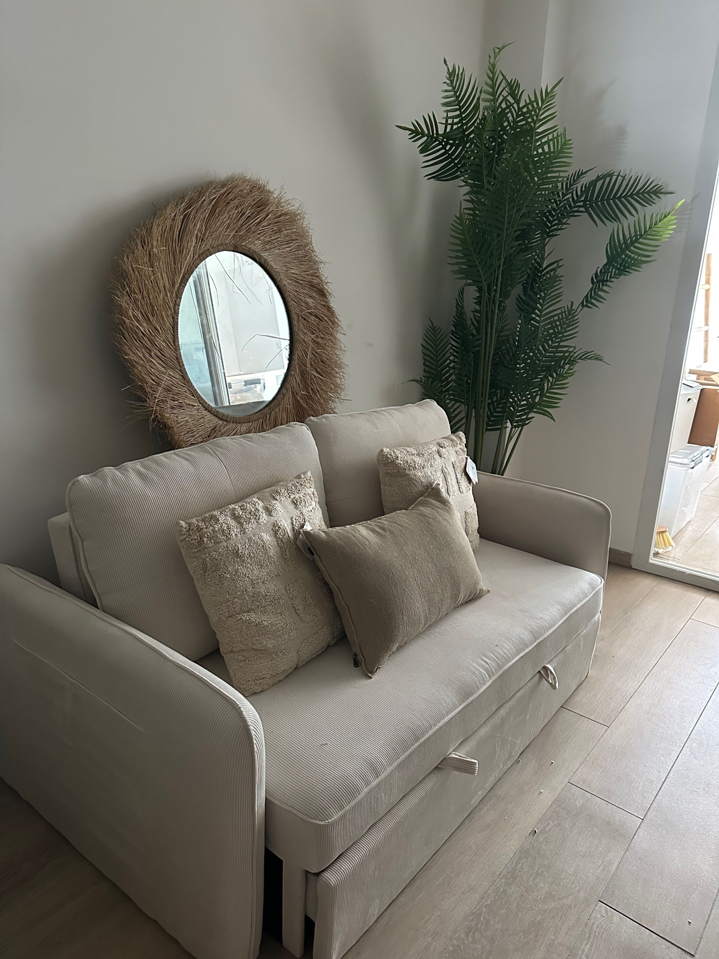 En beige soffa med två kuddar, en rund spegel med en ram i naturmaterial och en stor grön växt i ett hörn.