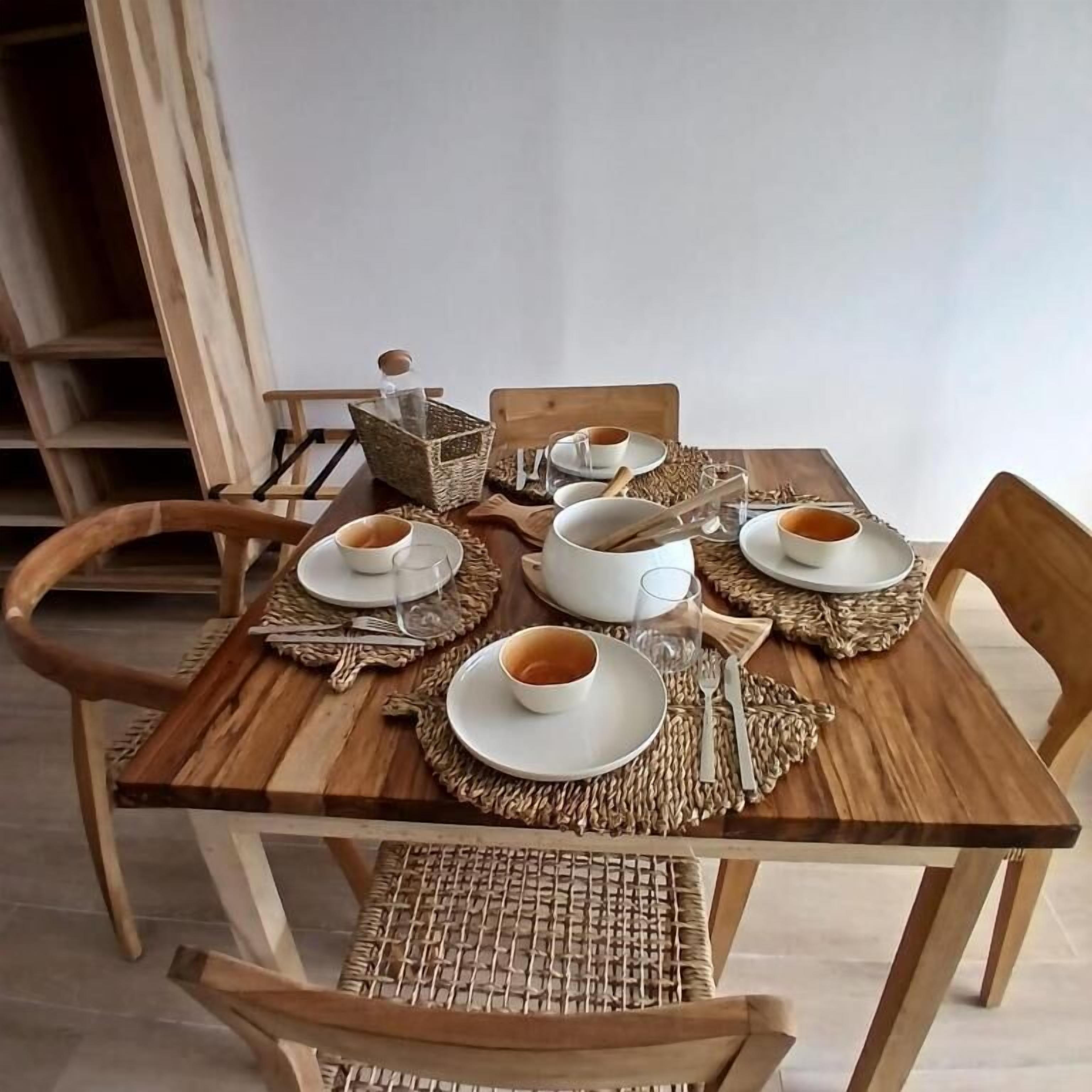 Una mesa de comedor de madera dispuesta con platos, cuencos y cubiertos blancos, rodeada de individuales tejidos.