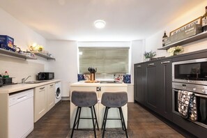 Een kleine keuken met een vaatwasser, magnetron en oven. Twee barkrukken en een raam met zonwering.
