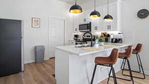 Una cocina moderna con armarios blancos, una isla central con taburetes, iluminación colgante, microondas y refrigerador.