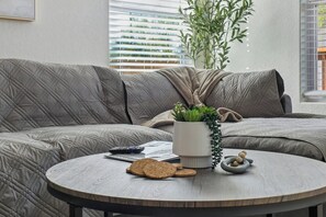 Un salon moderne avec un canapé gris à motifs matelassés, une table basse ronde en bois sur laquelle sont posés des biscuits et une petite plante, et une fenêtre avec des stores.