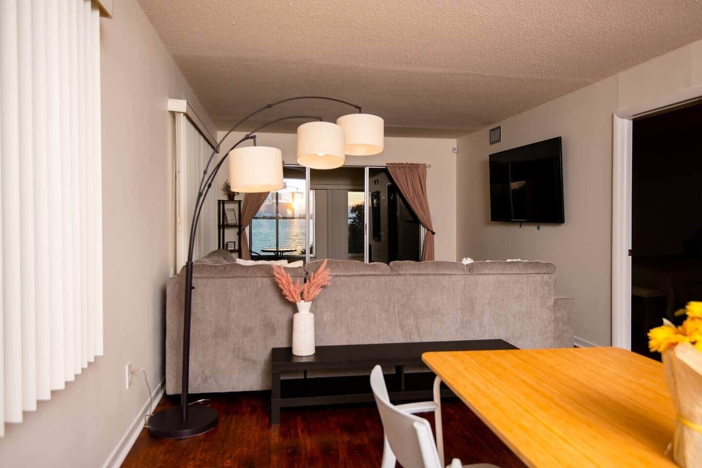 A modern living room with a sofa, a dining table, a floor lamp, and a TV mounted on the wall.