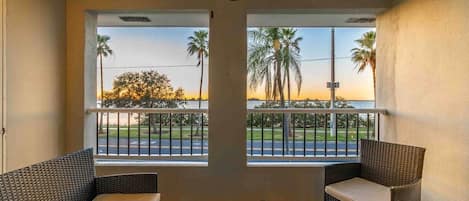 A balcony with wicker furniture, a ceiling fan, and a view of palm trees and a sunset.