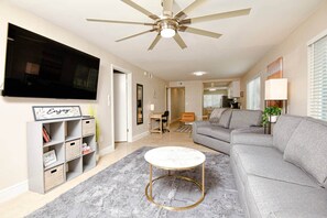 A modern living room with a grey sofa, a round coffee table, a ceiling fan, a TV mounted on the wall, and a bookshelf. - Waterfront Retreat 2BR 2BA (Clearwater)