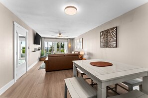 A modern living room with a sofa, a dining table with benches, a flat-screen TV mounted on the wall, and a ceiling fan. - Waterfront Condo in Dunedin/Clearwater (Clearwater)
