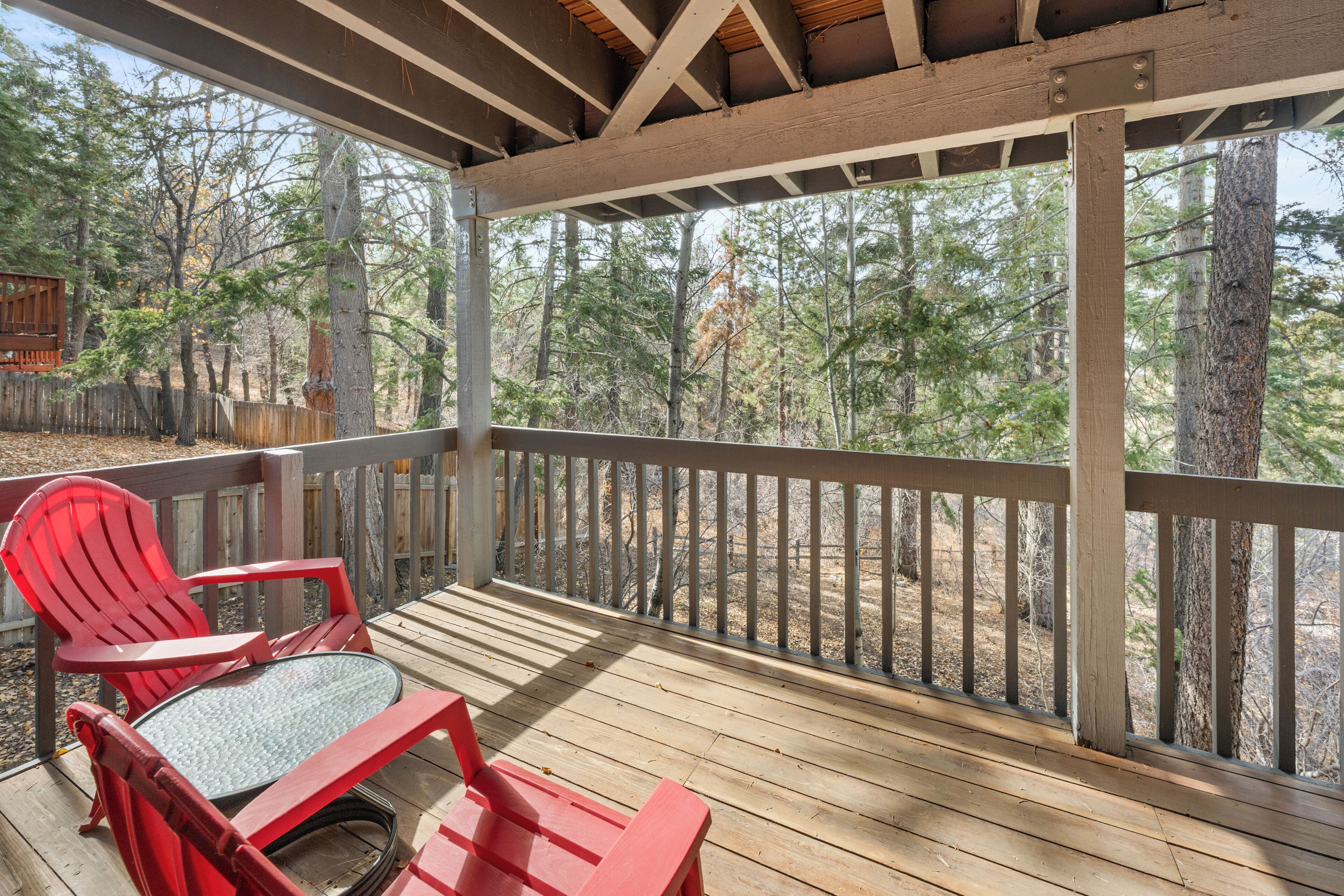 Una terraza de madera con una silla Adirondack roja y una mesa de vidrio, con vista a un área boscosa.