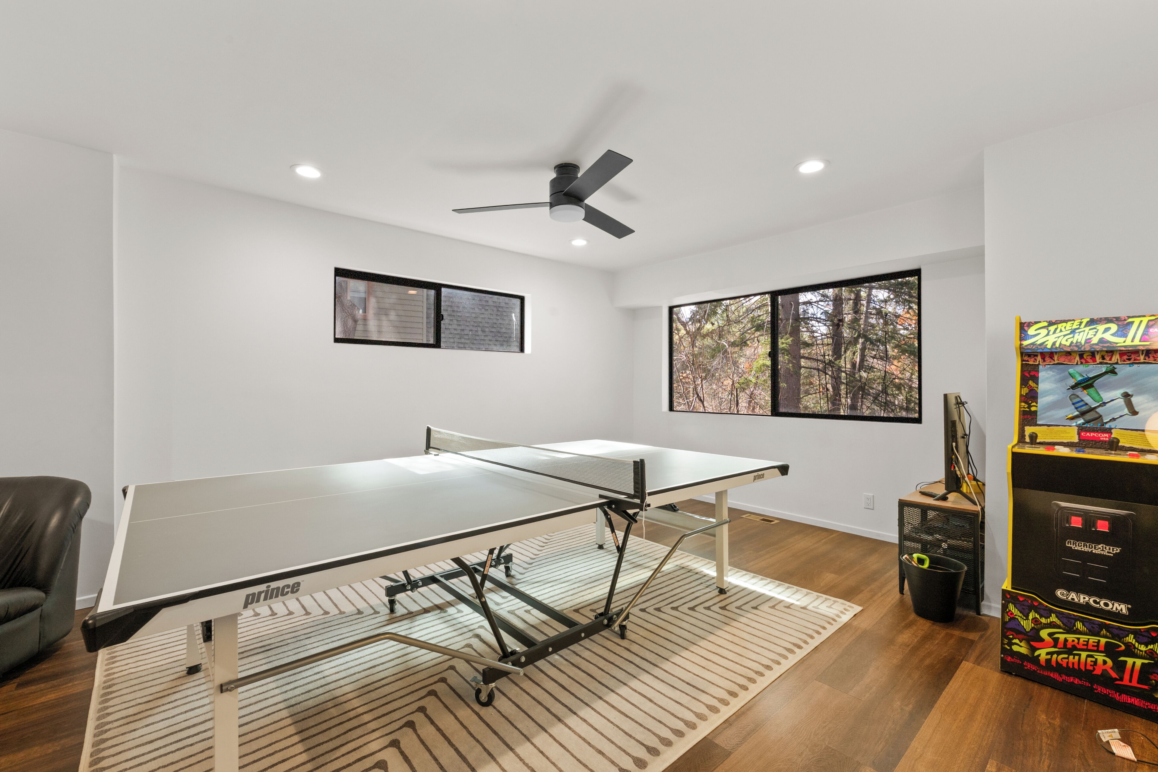A room with a ping pong table, ceiling fan, windows with forest views, and a Street Fighter II arcade machine.