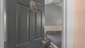 Une porte sombre fermée, ornée d’une poignée de porte en forme de lion, une fenêtre à cadre blanc et un lit avec une tête de lit blanche et une literie grise.