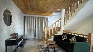 Una sala de estar con techo de madera, una escalera, un sofá negro, una mesa de centro redonda y una pequeña mesa con un espejo.
