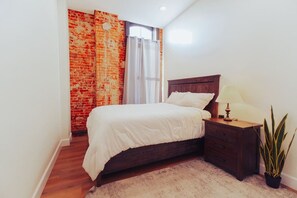Quarto com cama de madeira, mesa de cabeceira com abajur, planta em vaso e parede de tijolos.