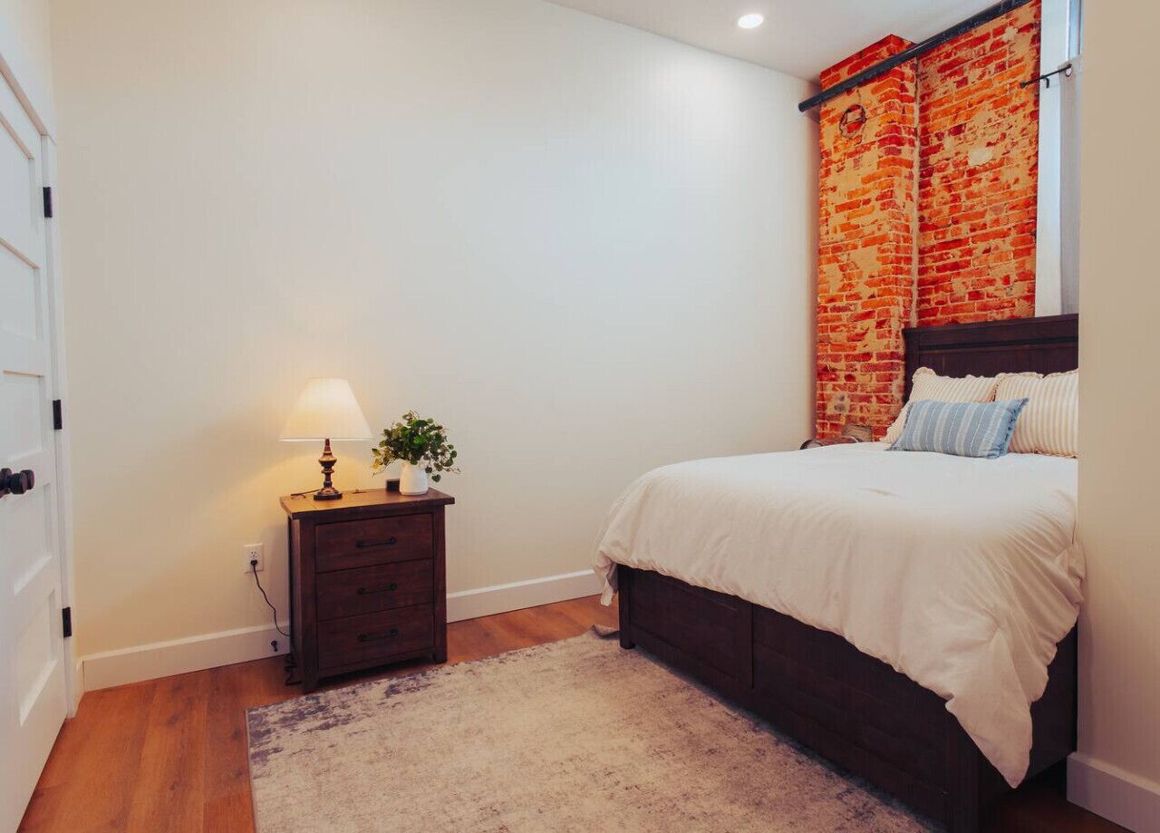 Een slaapkamer met een bed, nachtkastje met lamp, een dressoir en een gordijn met een baksteenpatroon.