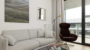 A modern living room with a grey sofa, a red armchair, a glass coffee table, and a large window with a view of buildings. - Oaza Spokoju | Widok na Las | Design | Shellter (Rogowo)