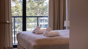 Ein Hotelzimmer mit Bett, zwei aufgerollten Handtüchern, einem Fenster mit Blick auf Bäume und einer Tür.