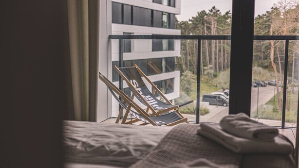Ein Balkon mit Stuhl und Handtüchern, im Hintergrund ein modernes Gebäude und Blick auf Bäume sowie eine Straße.