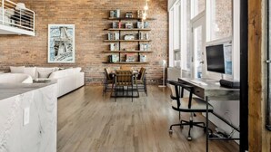 Un espace de vie moderne comprenant un mur en briques, un canapé blanc, un coin repas avec une table et des chaises, et un espace de travail avec un bureau et un ordinateur.