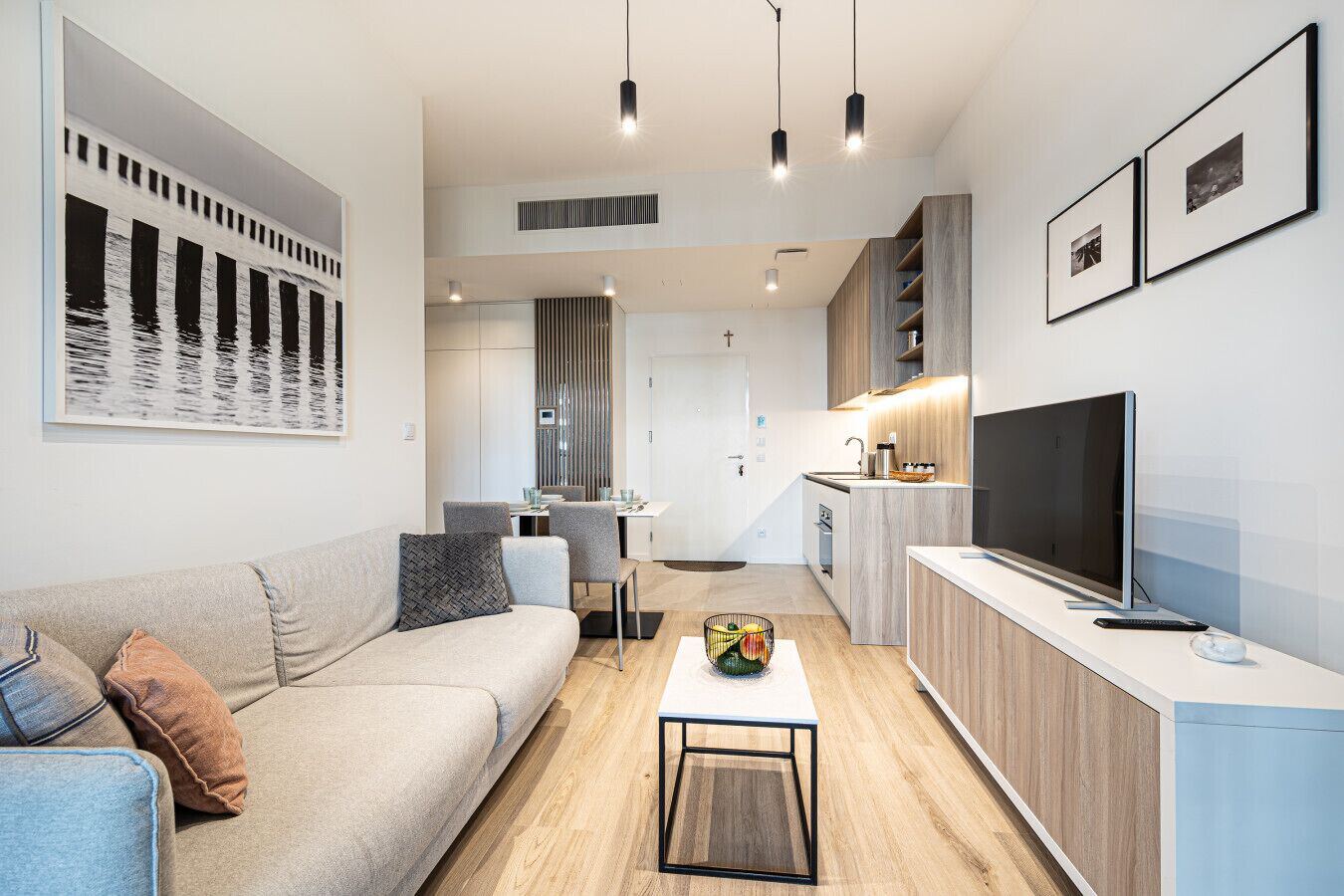 Ein modernes Wohnzimmer mit Sofa, Couchtisch und Fernseher. Es gibt eine Küchenzeile mit Holzschränken und einen Essbereich.
