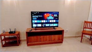 A television on a wooden stand with a remote control, a small table with a router, and a chair. -  Quartos Premium na 206 Sul  Banheiro Privativo (Brasília)