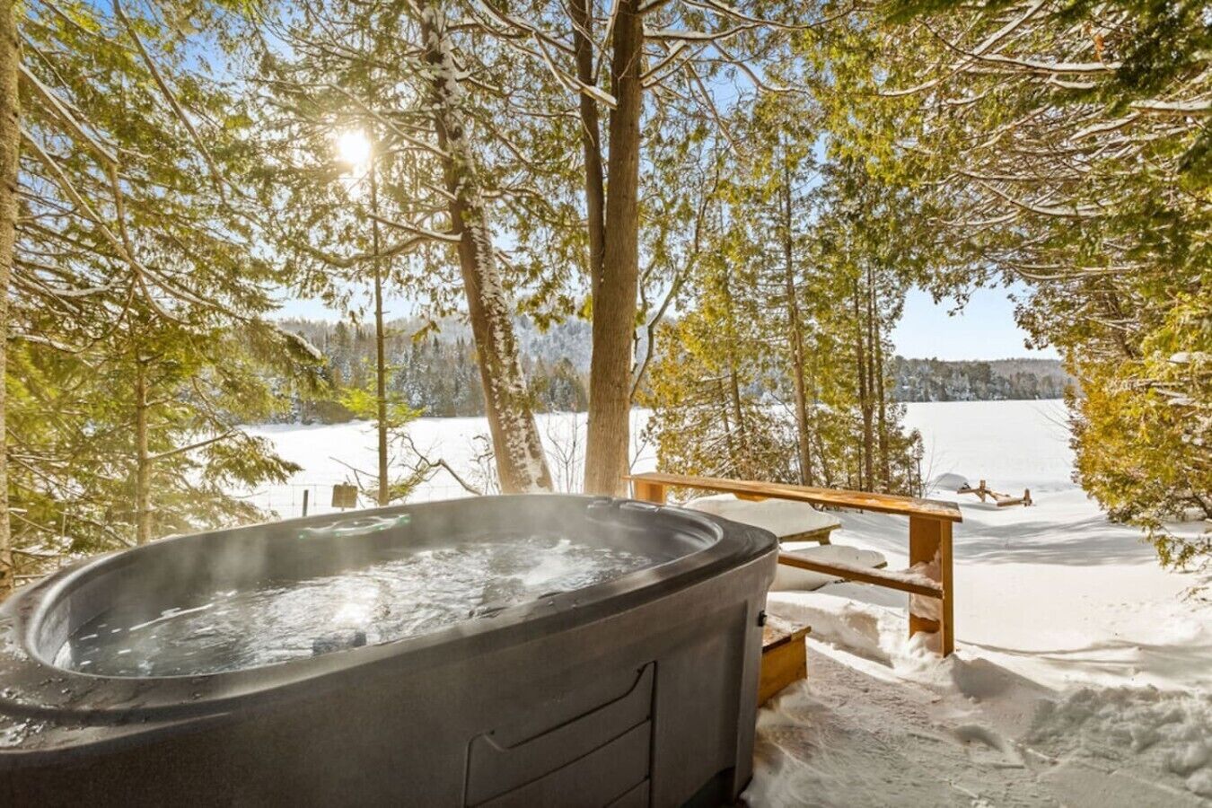 Uma banheira de hidromassagem em ambiente externo nevado, com vista para um lago e árvores.