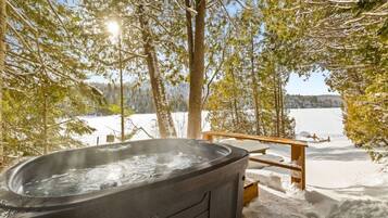 Uma banheira de hidromassagem em ambiente externo nevado, com vista para um lago e árvores.